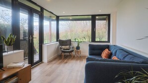 Ett modernt vardagsrum med en mörkblå soffa, ett soffbord i trä och en fåtölj med en kudde. Det finns stora fönster som ger utsikt över träd och en trädgård.