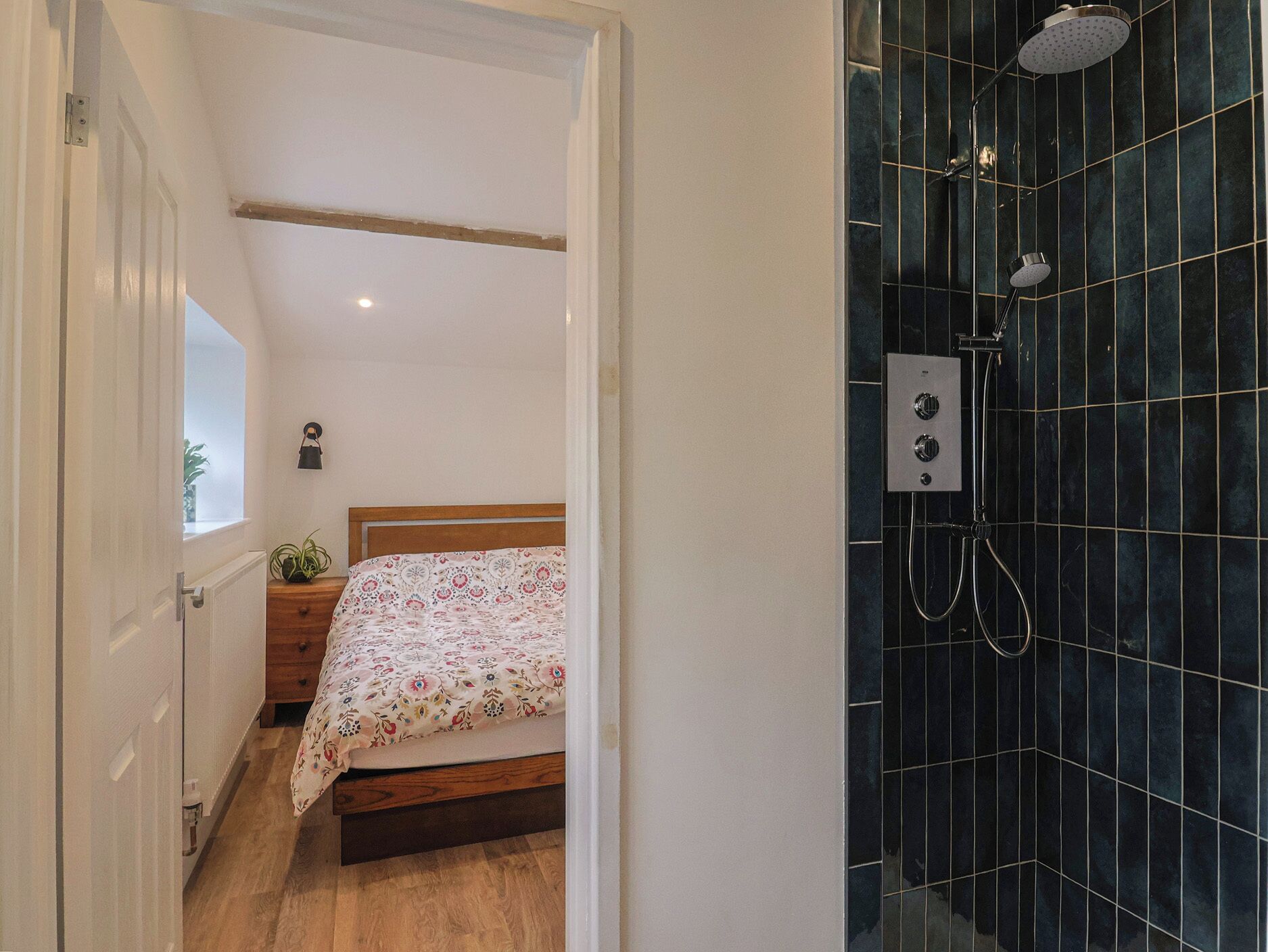 Ein Schlafzimmer mit einem hölzernen Bett, einem Nachttisch, einer kleinen Topfpflanze und einem Badezimmer mit dunkel gefliesten Wänden und einer Dusche.