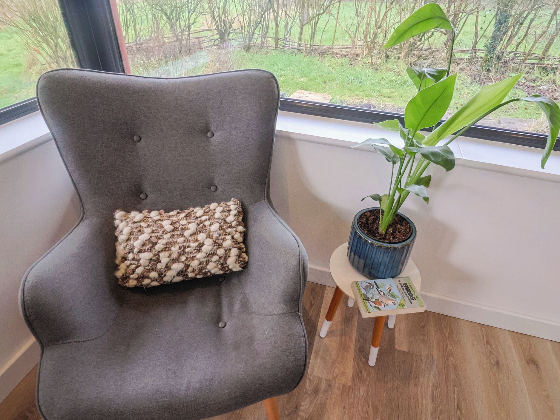 Un fauteuil gris recouvert de tissu, doté d’un coussin texturé, une petite table en bois sur laquelle est posée une plante en pot, et une fenêtre donnant sur un espace verdoyant.