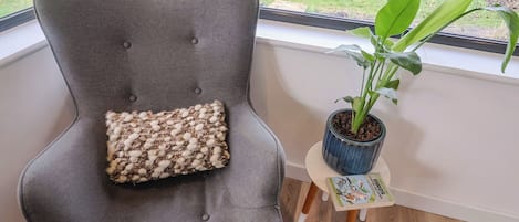 A grey upholstered chair with a textured cushion, a small wooden table with a potted plant, and a window with a view of a grassy area.
