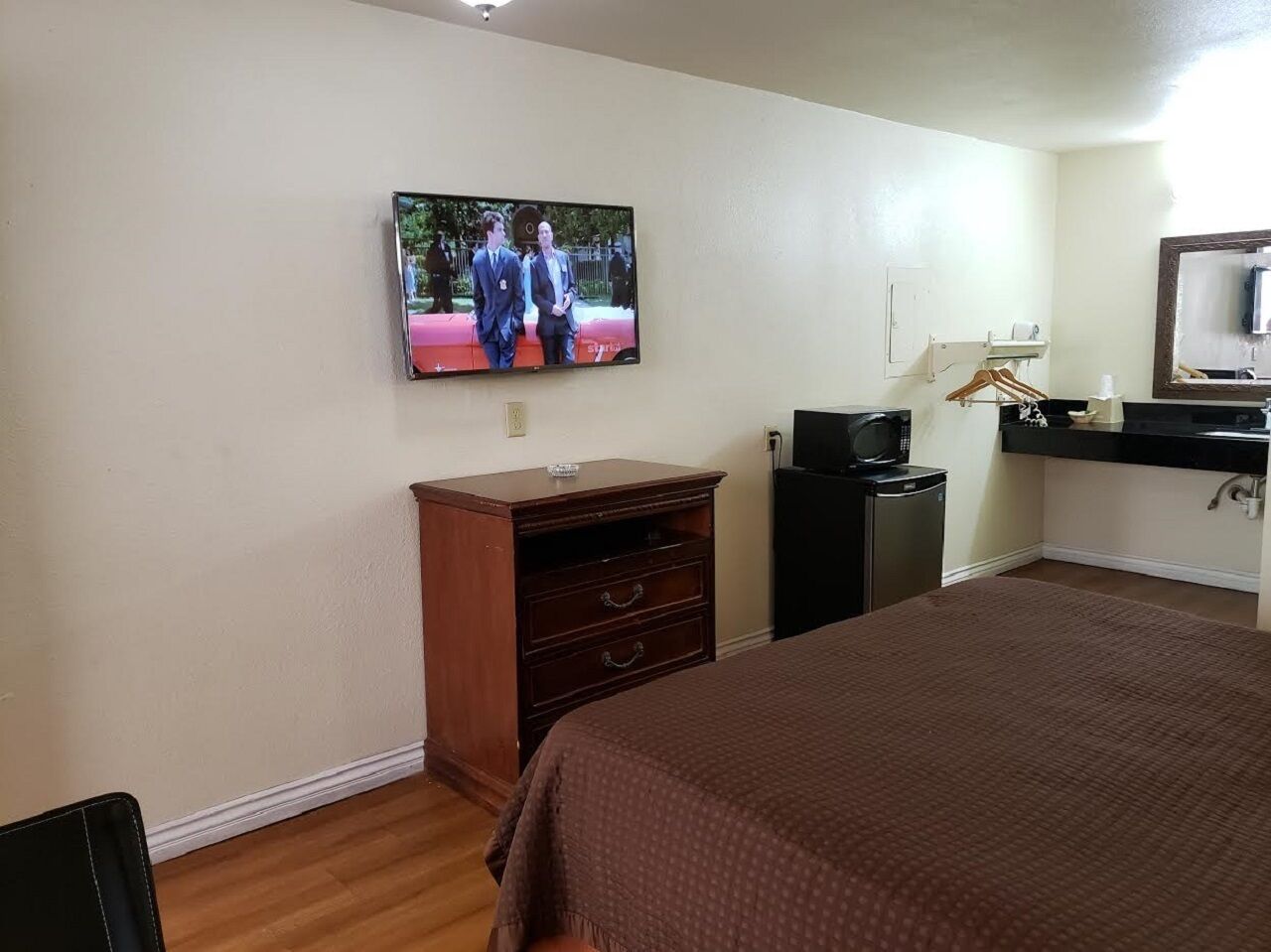 A hotel room with a bed, a TV mounted on the wall, a wooden dresser, a desk with a computer, and a mirror above the sink.