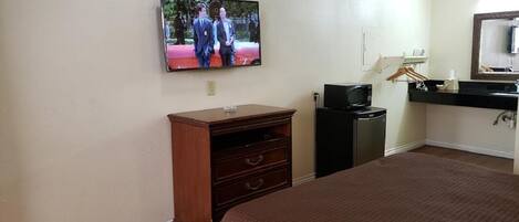 A hotel room with a bed, a TV mounted on the wall, a wooden dresser, a desk with a computer, and a mirror above the sink.