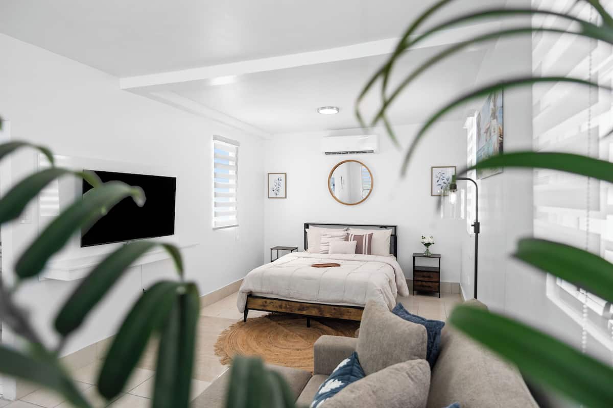 A bedroom with a bed, a TV, a bedside table, a mirror, and a window with blinds.