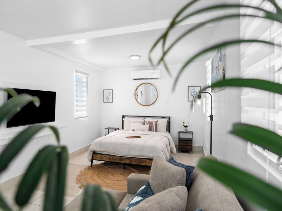 Un dormitorio con cama, televisor, mesita de noche, espejo y ventana con persianas.
