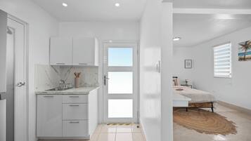 A modern kitchen with white cabinets, a marble countertop, and a view into a bedroom with a bed and a window.