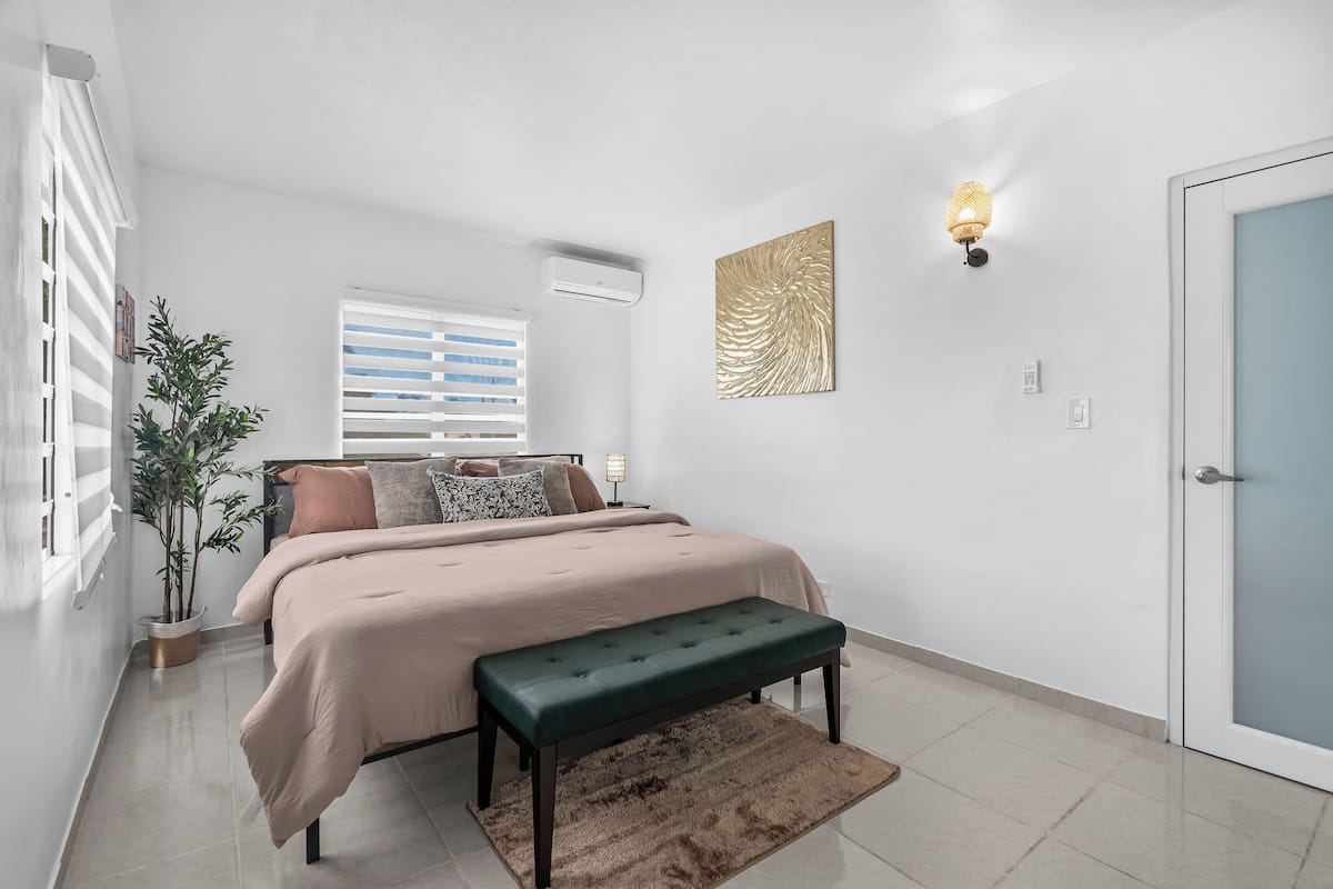 A bedroom with a bed, a bench, a potted plant, a wall-mounted light fixture, and a door.