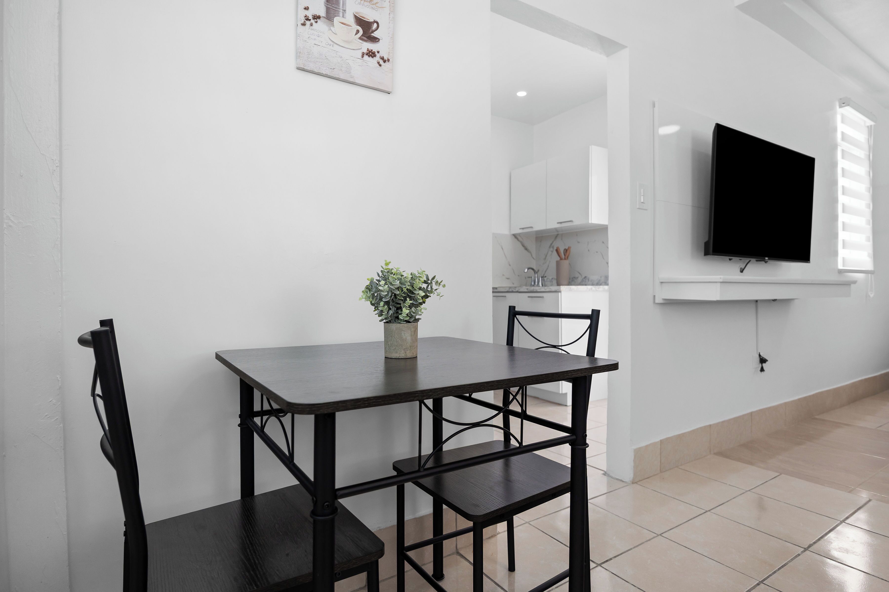 A modern dining area with a dark wooden table and chairs, a potted plant, a flat-screen TV mounted on the wall, and a kitchen area in the background.