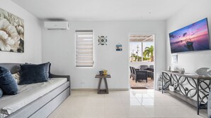 A modern living room with a grey sofa, a small table with snacks, a wall-mounted air conditioner, a decorative clock, and a TV displaying a sunset scene. - 63 Bay View by Otium (Catano)