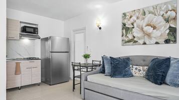 Una cocina moderna con microondas, estufa y refrigerador. Hay una mesa de comedor con sillas y un sofá gris con almohadas azules. Un cuadro grande con motivos florales adorna la pared.