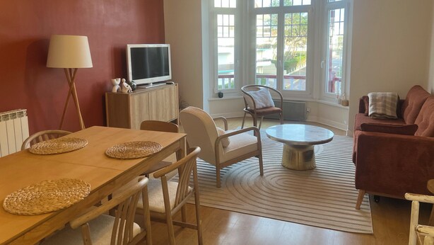 Un salón con una mesa de comedor de madera, sillas, un sofá, un televisor sobre un mueble bajo y ventanas amplias con vistas al exterior.