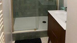 Una ducha con cerramiento de vidrio y marco dorado, un lavamanos blanco y una pequeña ventana.