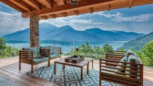 Une terrasse en bois avec un pilier en pierre, deux canapés en bois avec des coussins, une petite table et un tapis à motifs.