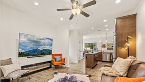 A modern living room with a fireplace, a large TV mounted on the wall, a ceiling fan, and a kitchen area in the background.