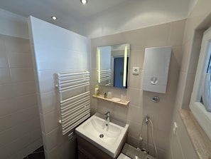 A modern bathroom with a white sink, a mirror, a towel rack, and a shower. - Apartments Benzia (Marina)