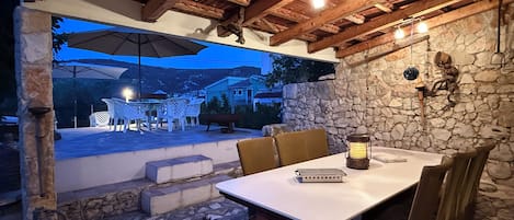 An outdoor patio with stone flooring, wooden beams, and a table with chairs. There is a stone wall and a view of houses in the background.