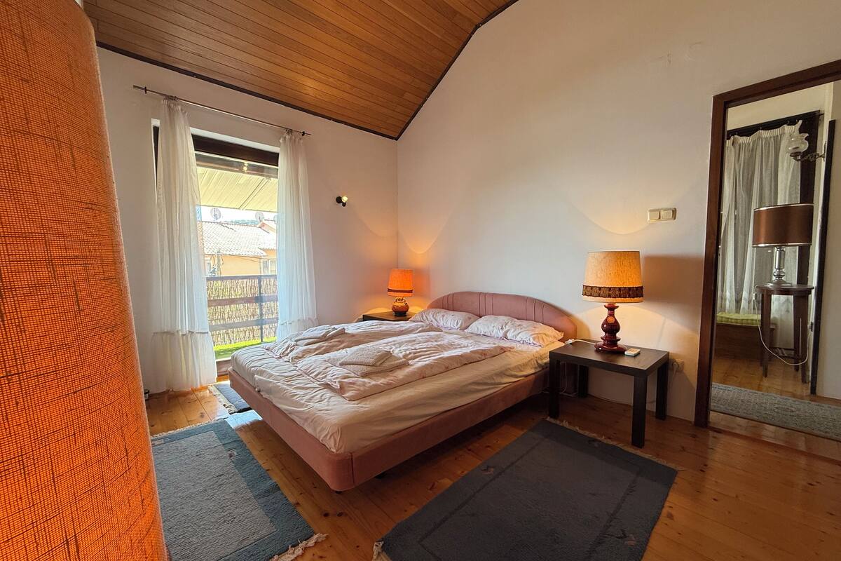 A bedroom with a bed, two bedside lamps, a wooden floor, and a window with curtains.