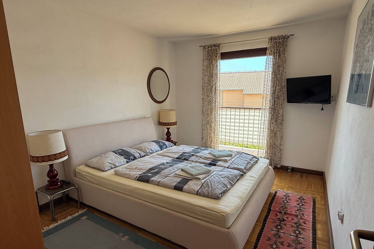 A bedroom with a bed, a nightstand, a lamp, a TV, and a window with curtains.