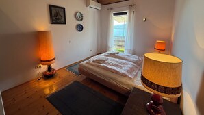 A bedroom with a bed, two lamps, a window with curtains, an air conditioner, and a small rug. - Apartments Benzia (Marina)