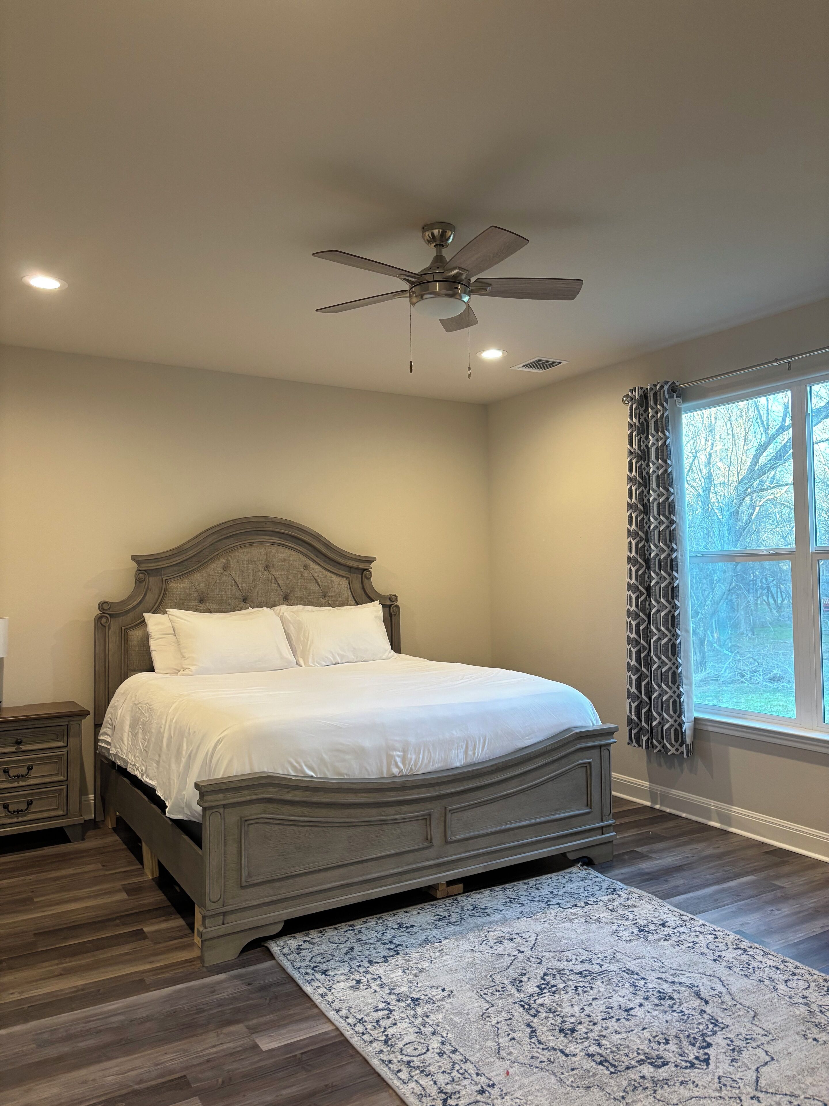 A bedroom with a bed, a ceiling fan, a window with curtains, and a nightstand.
