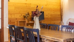 Een houten eettafel met zes stoelen, een vaas met gedroogde bloemen en een televisie op de achtergrond.