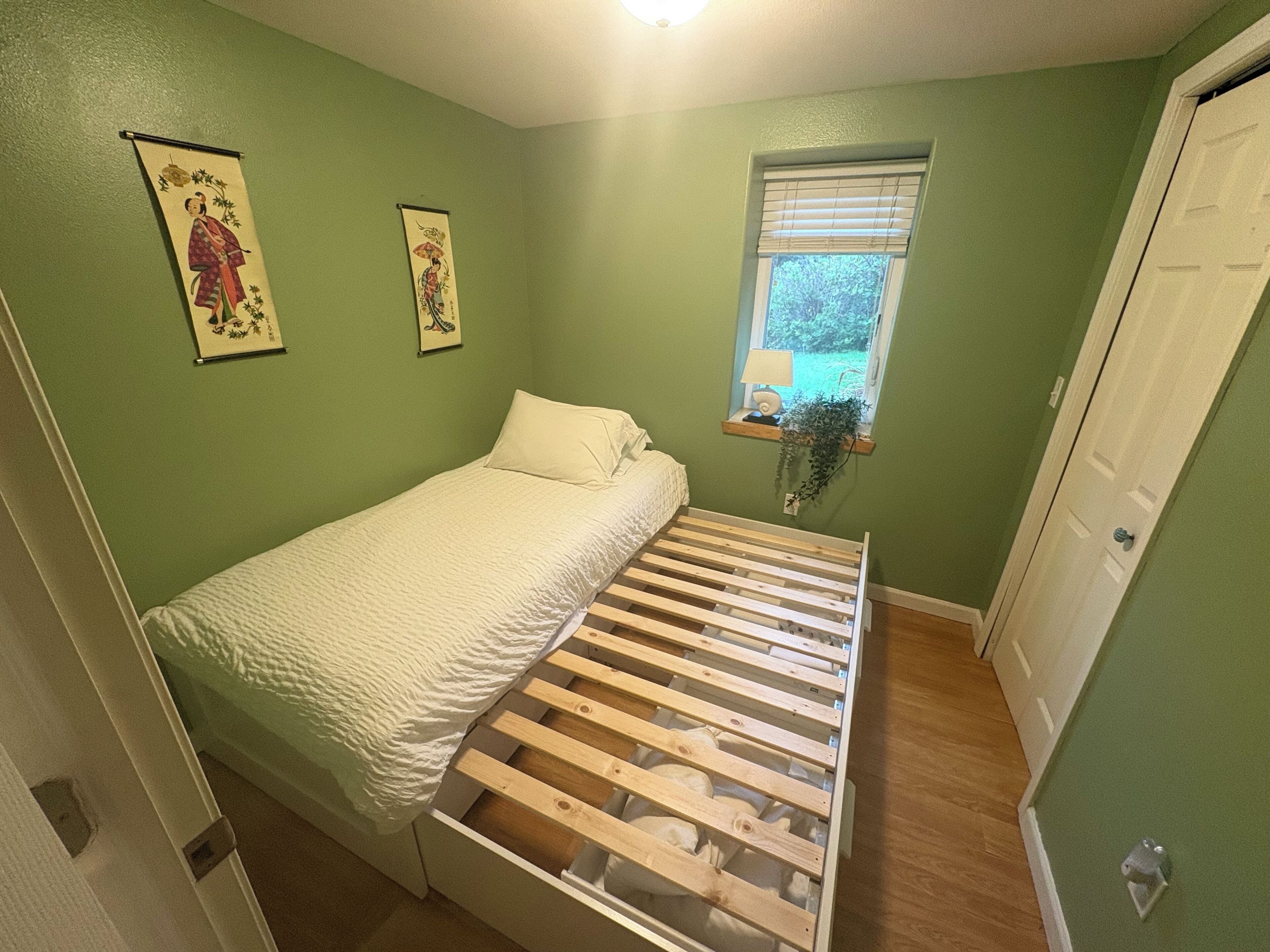 Una habitación pequeña con cama individual, cabecero de madera, paredes verdes y dos cuadros colgados.