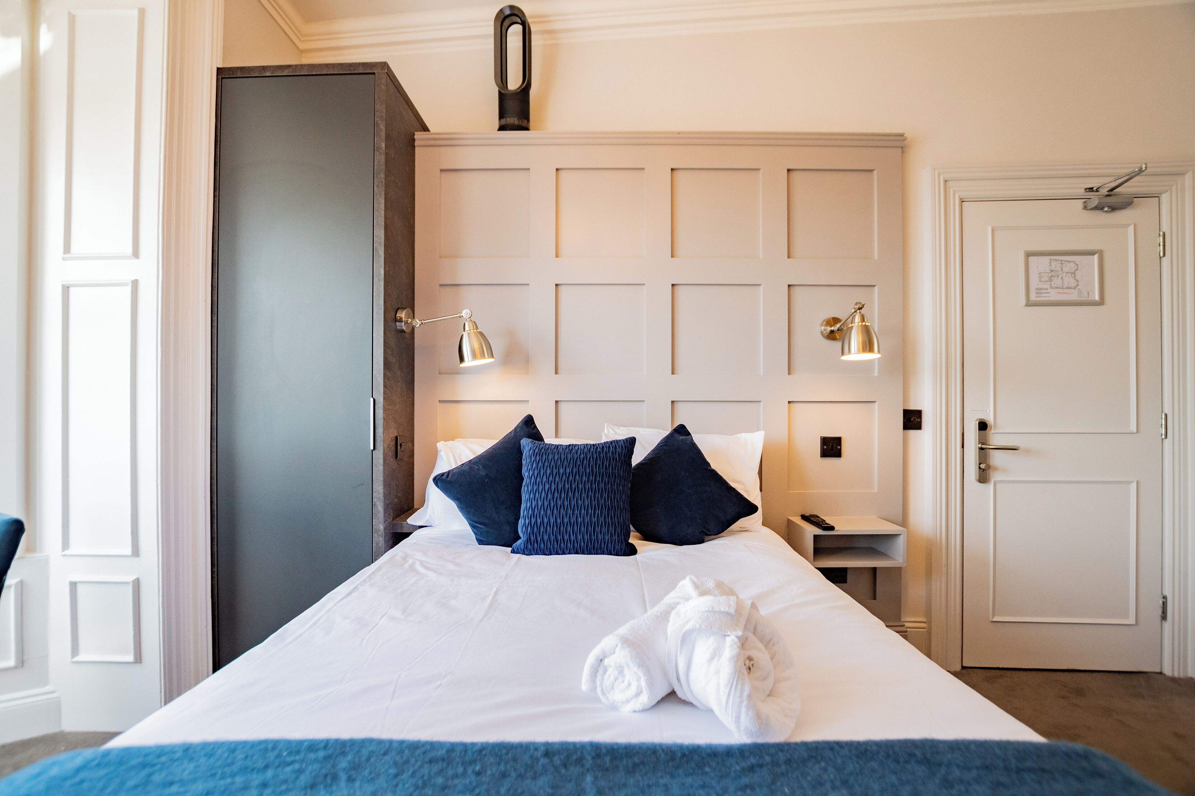A hotel room with a large bed, two bedside lamps, a white door, and a small table with a remote control.