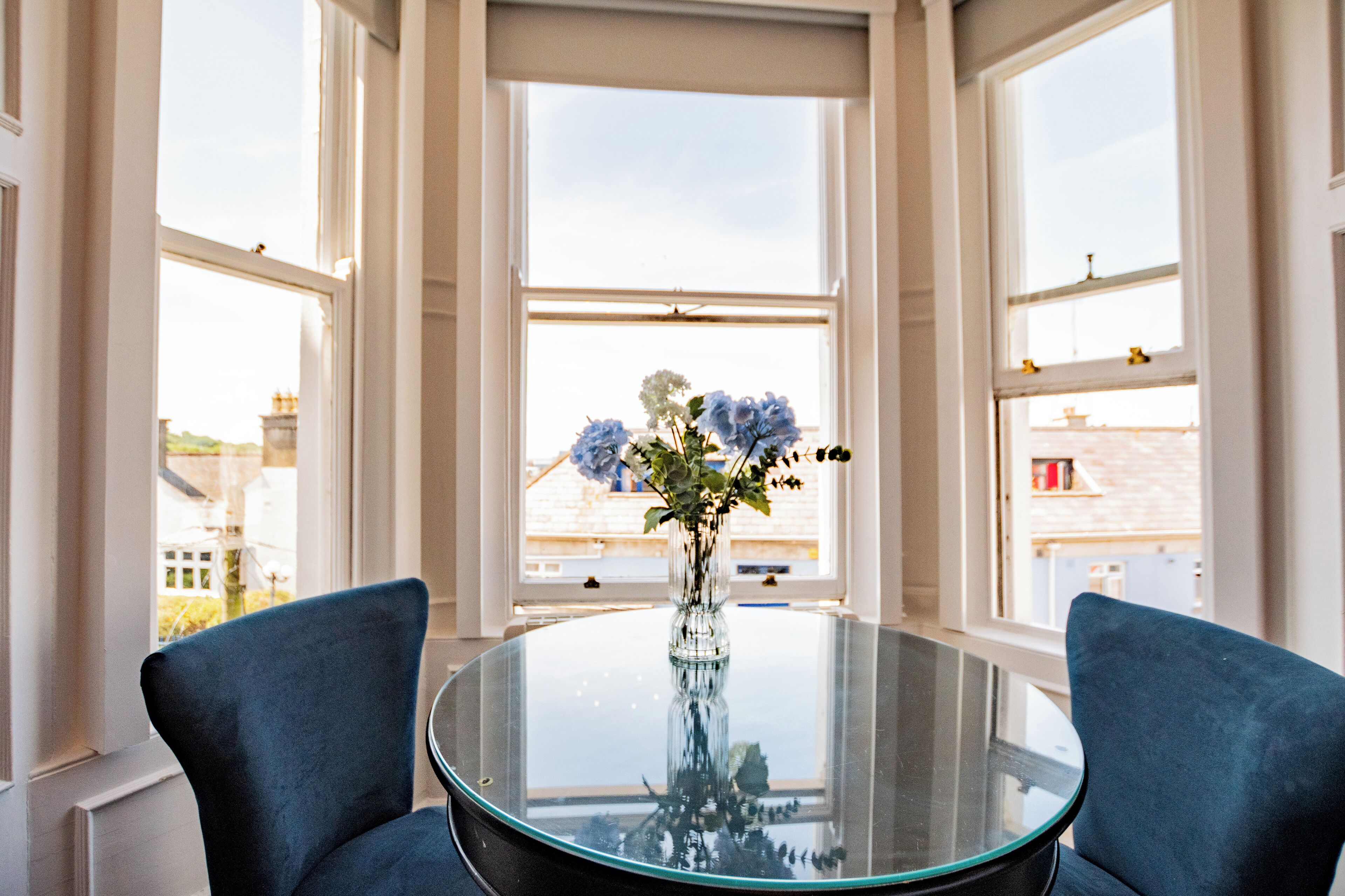 Una mesa de vidrio redonda con dos sillas azules, un florero con flores y vistas a edificios a través de amplios ventanales.