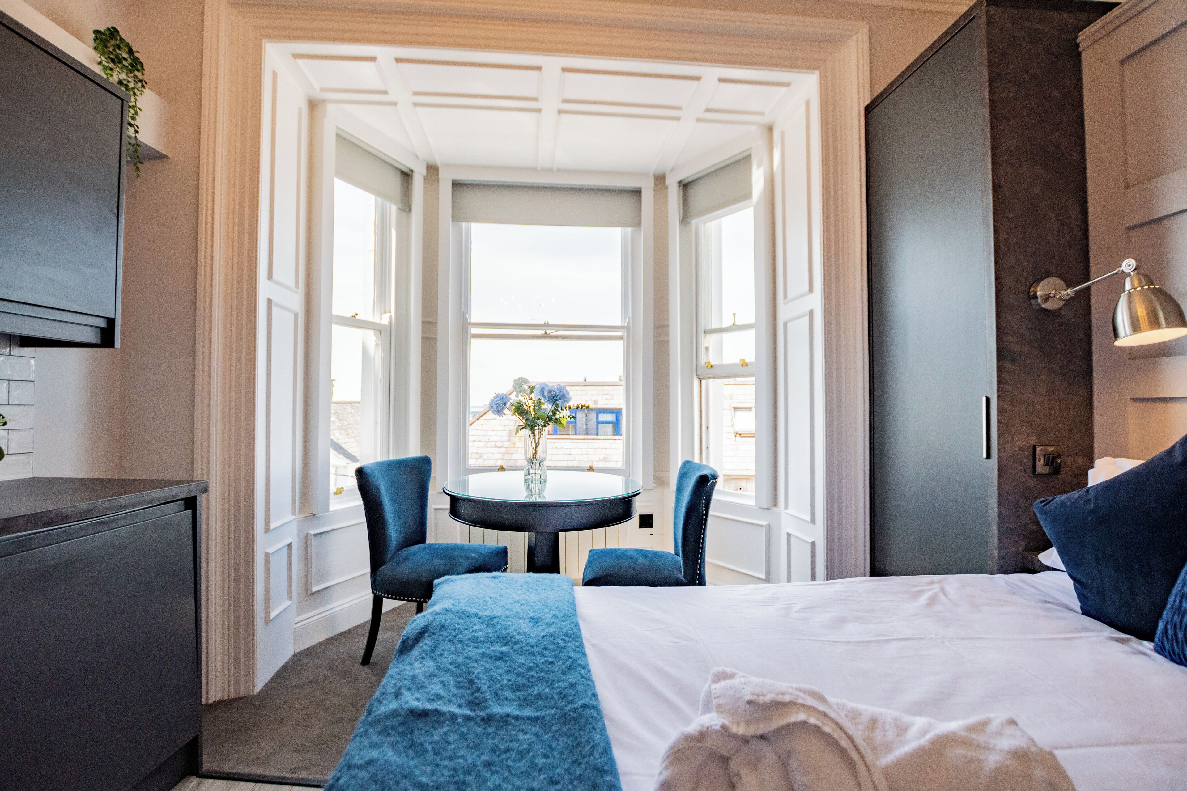A hotel room with a bed, a desk, a chair, a table with a vase, and a view through large windows.