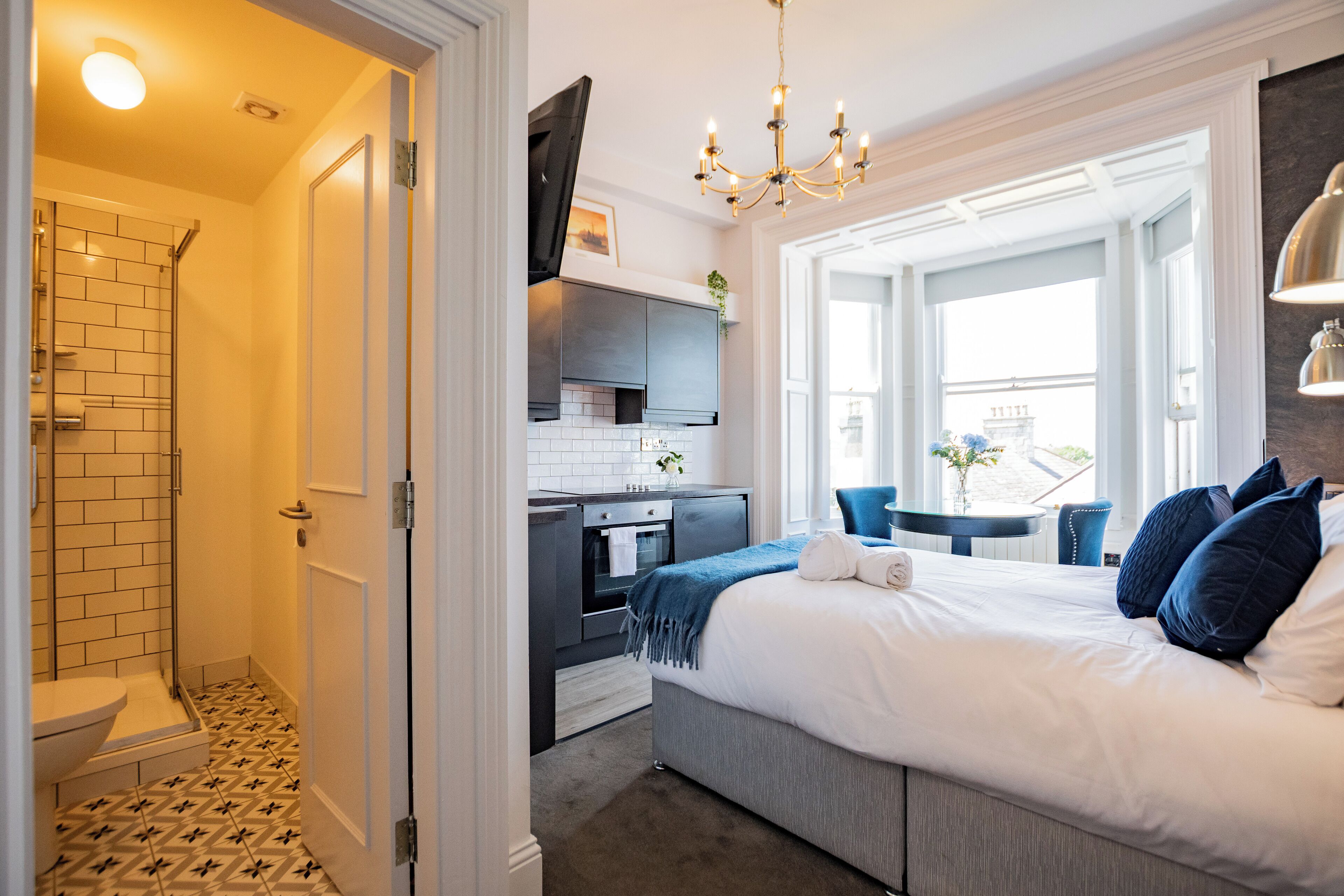 A modern hotel room with a bathroom, kitchenette, and a large bed with blue pillows.