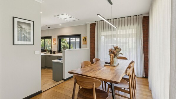 A modern dining area with a wooden table and chairs, a kitchen in the background, and a large window with curtains.