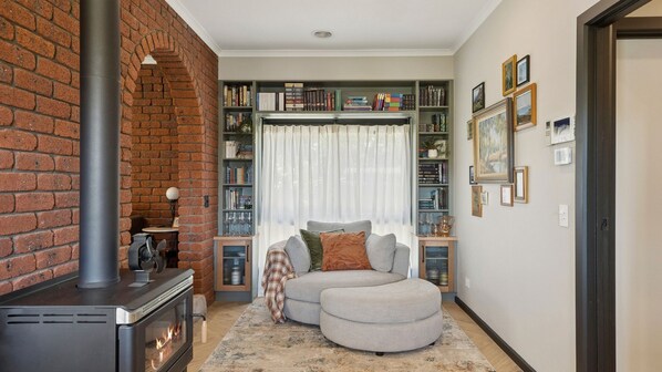 A cozy living room with a fireplace, a sectional sofa, a bookshelf, and a large window with curtains. - One Mile Creek Retreat (Wangaratta)