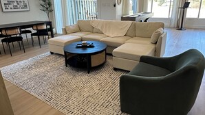Un salón espacioso con un amplio sofá modular, una mesa de centro redonda y un sillón oscuro. Se observa un comedor con una mesa y sillas, y un piano al fondo.