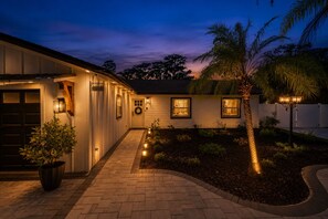 Una casa con un camino de ladrillo que lleva a la puerta principal, un letrero de bienvenida y un jardín con palmeras y plantas.