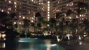 Ein Hotel bei Nacht mit mehreren Stockwerken, einem Schwimmbad und Palmen.