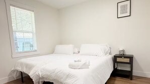 A bedroom with a large bed, a nightstand, a window with blinds, and a framed picture on the wall. - The Homestead House (Henderson)