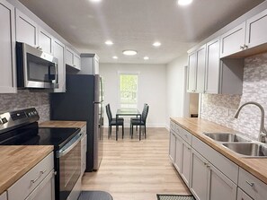A modern kitchen with a central island, built-in appliances, and a dining area with a window. - The Homestead House (Henderson)