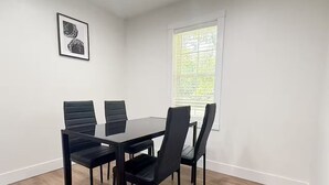A dining area with a black table and chairs, a framed picture on the wall, and a window with blinds. - The Homestead House (Henderson)