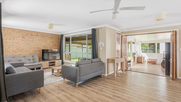 A modern living room with a brick wall, grey sofas, a TV, and a ceiling fan. - Boater's Retreat - pet friendly, right off the M1 (Boambee East)