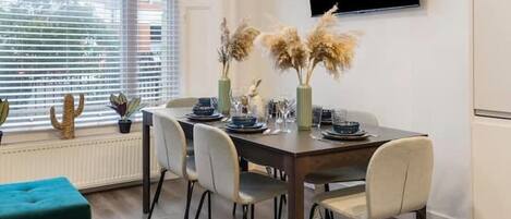 A dining area with a table set for four, chairs, a television mounted on the wall, and a window with blinds.