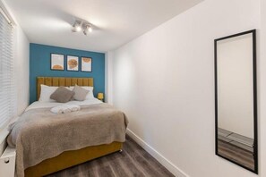 A bedroom with a bed, a headboard, a bedside table, a lamp, and a wall-mounted mirror. - Apartment - Sleeps 7 - Paddington Station 5 Mins (London)