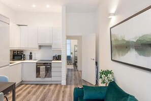 A modern kitchen with white cabinets, a microwave, and a stove. There is a dining area with a table and chairs, and a large window. - Apartment - Sleeps 7 - Paddington Station 5 Mins (London)