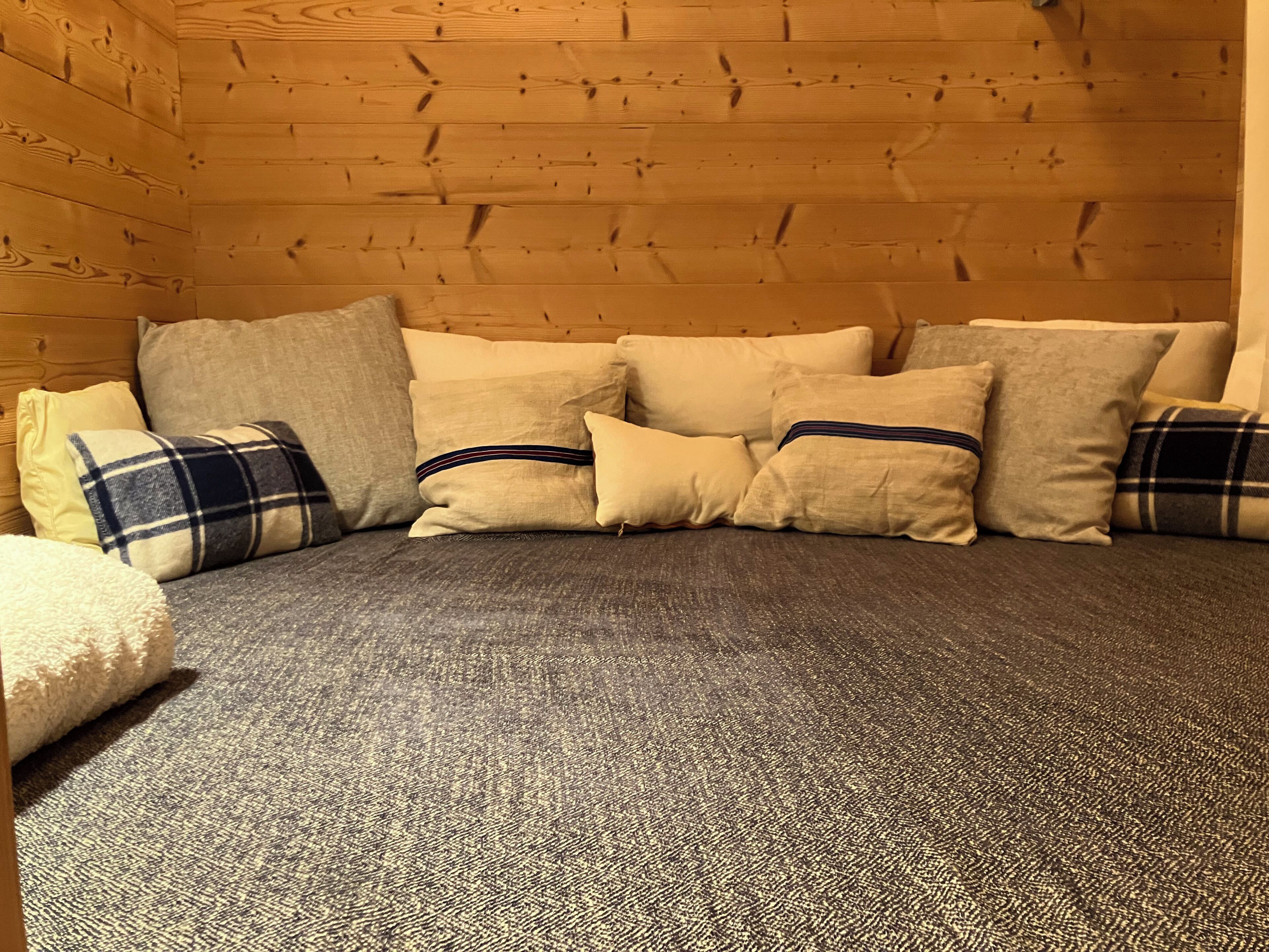 Ett mysigt rum med en grå soffa och flera kuddar, träväggar och ett golv täckt av en matta.