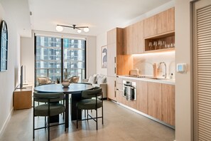 Een moderne keuken met houten kasten, een eethoek met een ronde tafel en stoelen, en een groot raam dat voor natuurlijk licht zorgt.