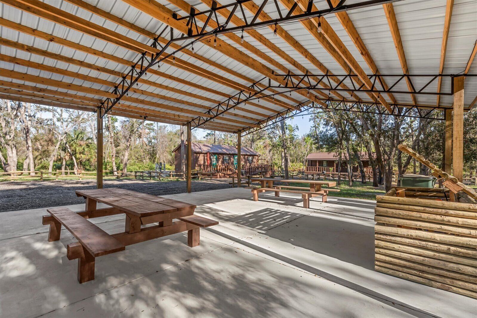 An outdoor covered picnic area with wooden benches and picnic tables, surrounded by trees and a rustic building in the background.