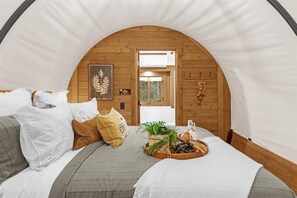 Una camera accogliente con soffitto in legno, un letto con biancheria bianca e grigia, un tavolino con una pianta e un boccale, e una porta che dà su una stanza luminosa.