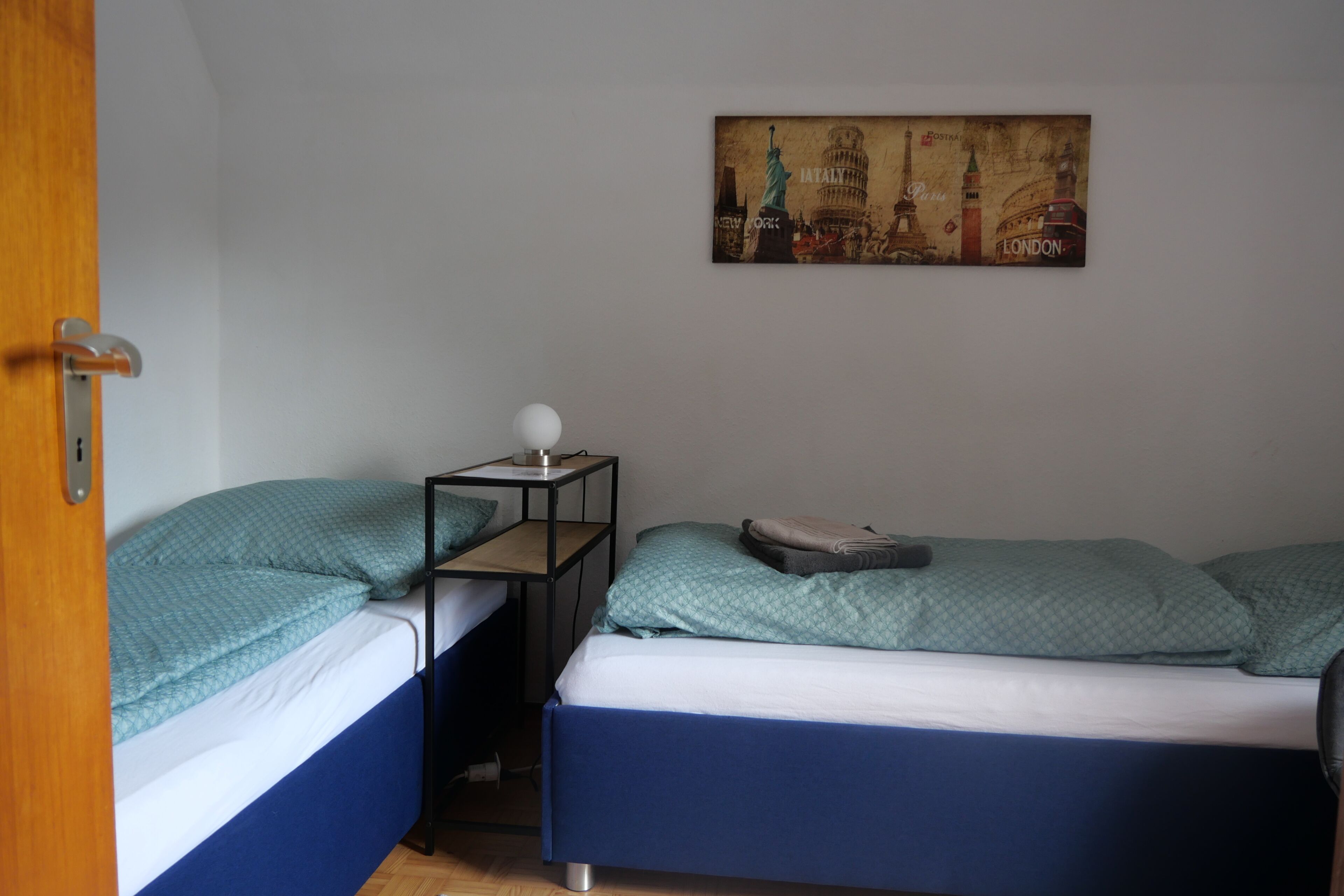 Ein Zimmer mit zwei Einzelbetten, einem kleinen Tisch mit Lampe und einem Wandbild bekannter Sehenswürdigkeiten.