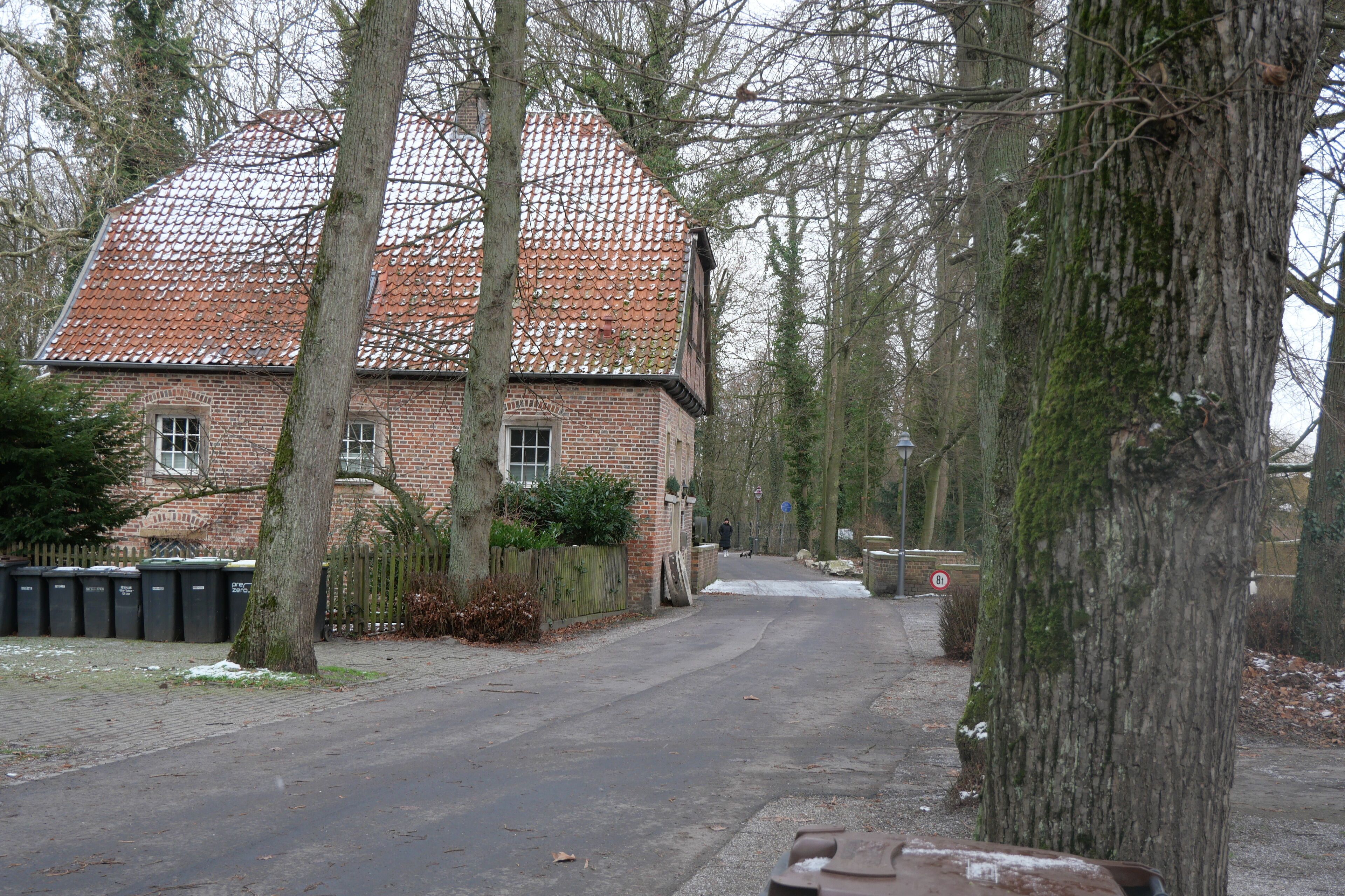 A brick building with a tiled roof, surrounded by trees and a paved road.