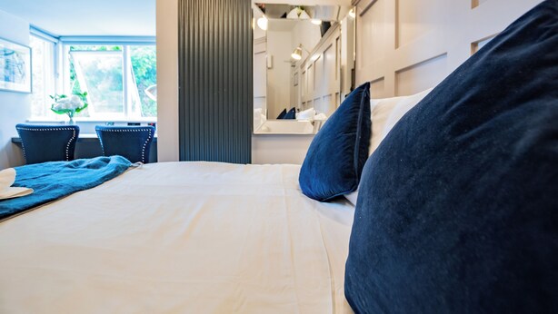 A modern hotel room with a large bed, a sitting area with chairs, a window with a view of greenery, and a mirror above the bed. - Modern Studio by Howth Marina (Howth)