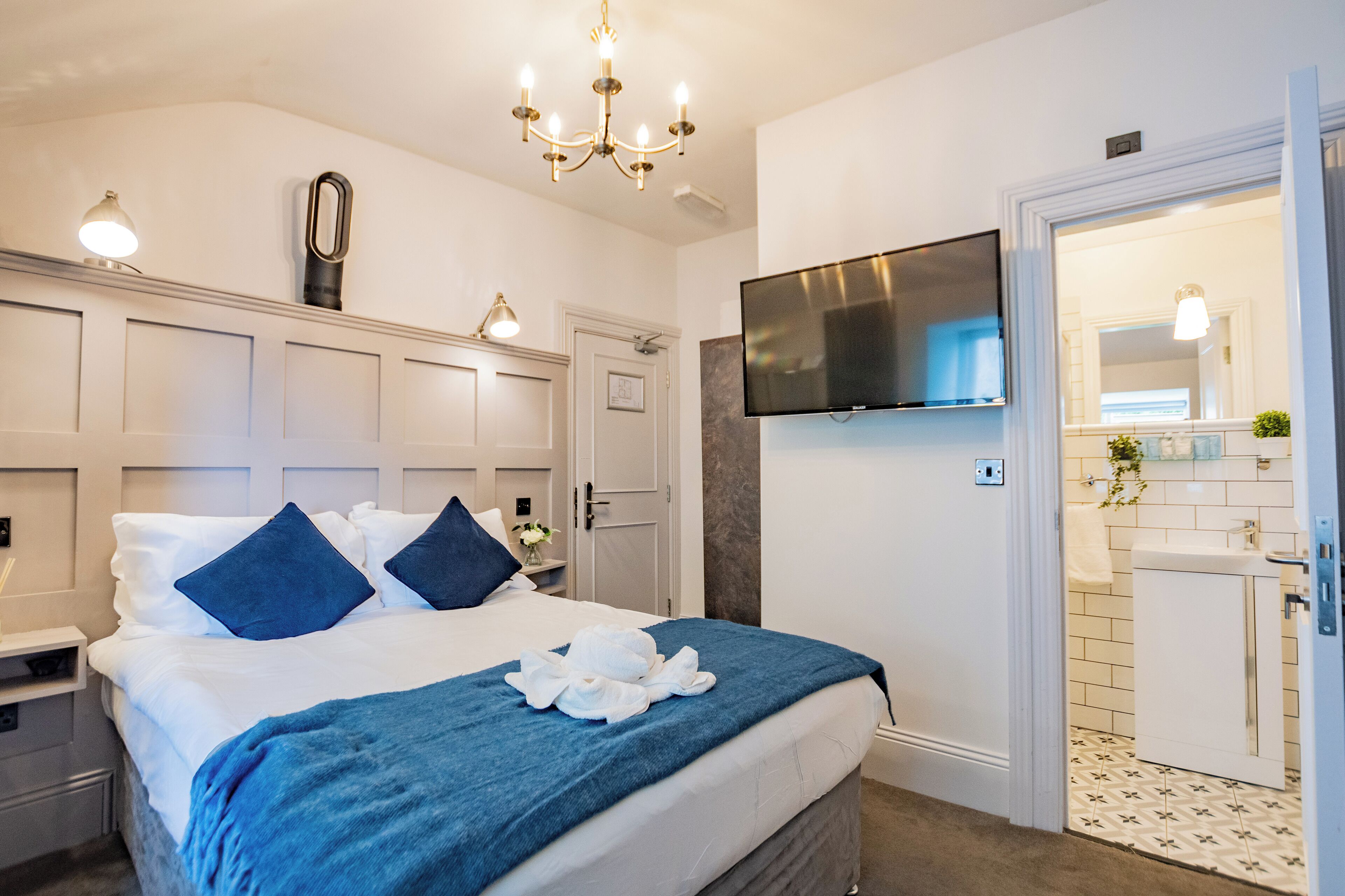 Habitación de hotel con cama, televisor montado en la pared, baño con lavamanos y espejo, y un candelabro.
