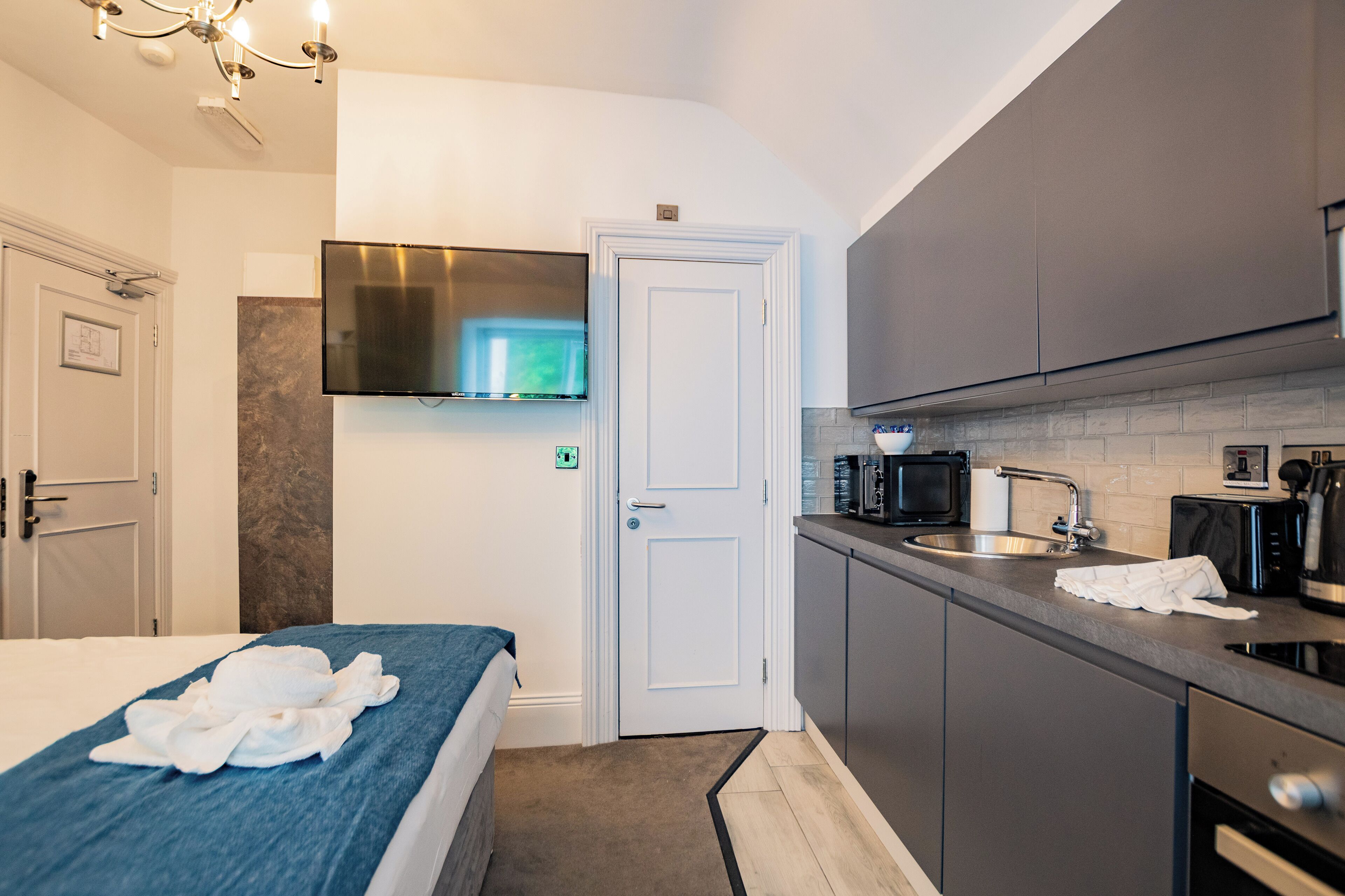 Hotellihuone, jossa on sänky, seinälle asennettu taulutelevisio oven yläpuolella, pieni keittiö, jossa on mikroaaltouuni, leivänpaahdin ja pesuallas, sekä kattokruunu.